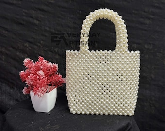 Puede incluir: Un bolso blanco con cuentas de forma rectangular y un asa redondeada. El bolso está hecho de pequeñas cuentas redondas. A la izquierda del bolso hay una pequeña maceta blanca con flores rojas. El texto "EVERYDAY ARTISAN" es visible.