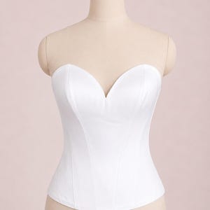 May include: A strapless white corset top featuring a sweetheart neckline and vertical seam detailing. The garment is constructed with a structured bodice that tapers at the waist, providing a fitted silhouette. The fabric is a solid, opaque white.