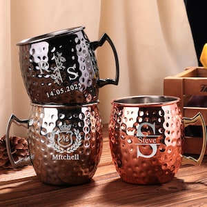 Pode incluir: Três canecas Moscow Mule em aço inoxidável com acabamento martelado. Uma caneca preta tem a letra "S" e a data "14.05.2025". As outras canecas são de cobre e prata com iniciais e nomes personalizados.