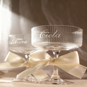 May include: Two clear glass champagne coupe glasses with ribbed detailing and the names "Ciela" and "Aime" etched on them. Each glass has a cream-coloured ribbon tied around the stem. The background is softly lit.