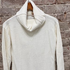 May include: Off-white knit sweater with a cowl neck and long sleeves. The sweater is made of a textured knit fabric and hangs on a wooden hanger against a brick wall background. A casual, comfortable top.