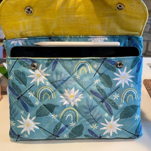 May include: A quilted tablet case with a teal background featuring dragonflies, white flowers, and rainbows. The interior is yellow. The case is open, revealing a tablet and stylus inside.