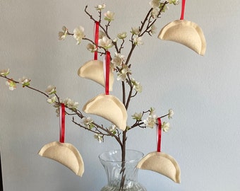 Può includere: Un vaso bianco pieno di perline di vetro trasparente si trova su un runner da tavolo blu. Un ramo con fiori bianchi è decorato con sei ornamenti in feltro a forma di mezzaluna, ognuno con un nastro rosso.