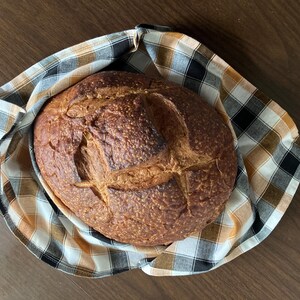 May include: A round loaf of freshly baked bread with a scored top, resting in a checkered cloth. The bread has a golden-brown crust and is surrounded by a fabric with a black, white, and tan plaid pattern. The image is taken from an overhead perspective.