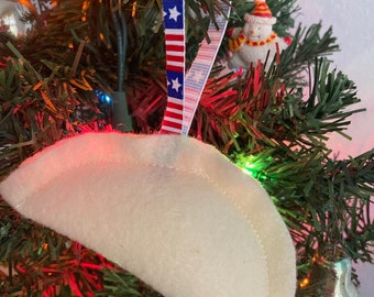 May include: A white felt ornament shaped like a half-moon with a red, white, and blue ribbon attached. The ornament is hanging on a Christmas tree.