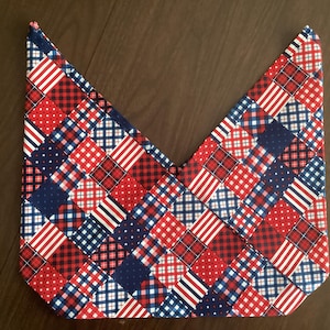 May include: A quilted fabric item with a patchwork design in red, white, and blue. The pattern includes stripes, polka dots, and plaid squares, creating a patriotic theme. The item has a unique shape.
