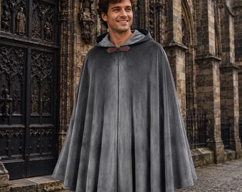 May include: A gray velvet hooded cloak with a brown leather clasp. The cloak is worn by a person standing in front of a stone building with ornate doors. The cloak has a flowing design.