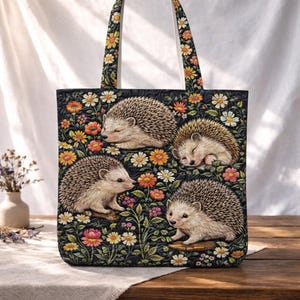 Könnte beinhalten: Eine schwarze Tragetasche mit einem Blumen- und Igel-Design. Die Tasche zeigt vier Igel, die von bunten Blumen umgeben sind. Die Griffe sind mit Blumen gemustert. Die Igel sind realistisch dargestellt. Die Blumen umfassen Gänseblümchen und andere Blüten.