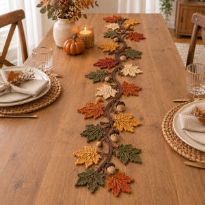 Puede incluir: Un camino de mesa de ganchillo que presenta una larga enredadera marrón sinuosa adornada con varias hojas de colores otoñales en tonos naranja, amarillo, verde oscuro y crema, intercaladas con pequeñas bellotas de ganchillo de color marrón claro.