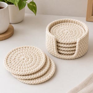 May include: A set of round, cream-coloured crocheted coasters made from thick yarn. The set includes a matching circular holder with a U-shaped cutout, containing a stack of coasters, while three additional coasters are arranged in an overlapping row.