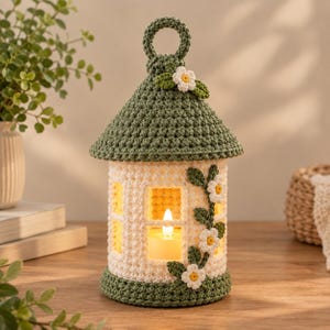 May include: A crocheted lantern featuring a cream-colored cylindrical body with window cutouts and a conical olive green roof topped with a circular handle. The structure is decorated with small white flowers and green leaves.