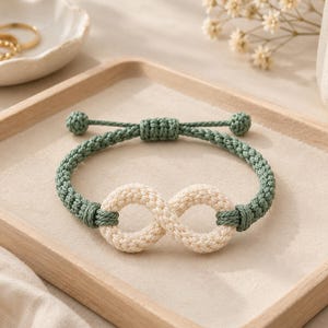 May include: A cord bracelet featuring a central infinity symbol crafted from cream-colored woven rope, connected to a sage green braided band. The bracelet includes an adjustable sliding knot closure with two small knotted ends for a secure fit.