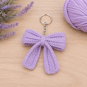 May include: A light purple crochet bow keychain featuring a textured, ribbed yarn construction. The bow is attached to a silver-toned metal split ring by a short chain link, showcasing a consistent lavender color throughout the handmade fiber design.