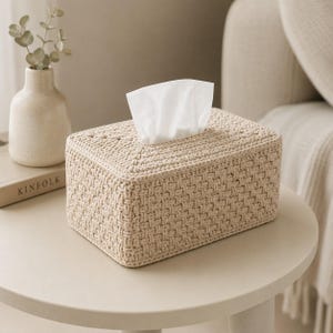 May include: A rectangular tissue box cover crafted from beige yarn in a textured basketweave pattern. The cover features a central opening on the top surface, through which a single white tissue is pulled upward, displaying a clean and neutral aesthetic.