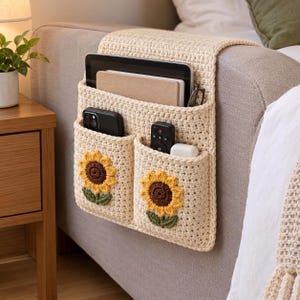 May include: A cream-coloured crochet sofa armrest organiser featuring two front pockets, each decorated with a sunflower motif. The organiser holds a tablet, a notebook, a smartphone, a remote control, and a small white case in its compartments.
