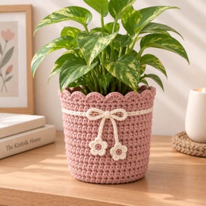 May include: A dusty pink crocheted plant pot cover featuring a scalloped top edge and a white crocheted ribbon tied in a bow. Two small white crocheted flowers with pink centres hang from the bow. A green and yellow variegated pothos plant sits inside.
