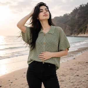 May include: A sage green short-sleeved knit shirt featuring a collared neckline, a button-down front with three visible buttons, and a textured open-stitch pattern. The garment is paired with solid black pants, creating a simple and clean look.