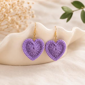Può includere: Un paio di orecchini pendenti con ciondoli a forma di cuore realizzati all'uncinetto in color lavanda. Ogni cuore è sospeso a un gancio in metallo dorato, mostrando una trama di filato dettagliata e una tonalità viola vivace e uniforme.