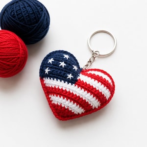 May include: A heart-shaped crochet keychain featuring the red, white, and blue pattern of the American flag, with a dark blue corner containing white embroidered stars and alternating red and white stripes, attached to a silver metal keyring.