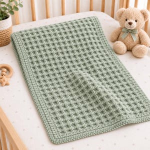 Puede incluir: Una manta rectangular de ganchillo en color verde salvia con un patrón de textura de gofre y un borde trenzado grueso. La manta se muestra junto a un oso de peluche marrón claro con un lazo de cinta verde en el cuello.