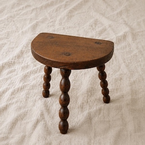 Puede incluir: Un taburete de madera pequeño en forma de semicírculo con un acabado marrón oscuro y tres patas. Cada pata está construida con una serie de segmentos apilados, redondeados y similares a cuentas. El asiento plano muestra vetas de madera.