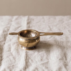 Pode incluir: Um coador de chá de latão polido com uma taça circular de base perfurada e um cabo longo e plano. O coador tem uma borda larga e uma pequena base circular de pedestal, apresentando um acabamento metálico dourado com gravuras.