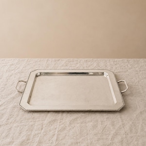 May include: A rectangular silver-toned serving tray featuring a smooth, flat centre surface and a wide, angled border with a beaded edge detail. The tray includes two small, angular metal handles attached to the shorter sides of the frame.