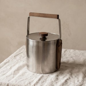 May include: A cylindrical stainless steel ice bucket with a brushed metal finish. It features a dark wood handle attached to a metal swing arm and a matching dark wood knob on the lid. The design includes vertical wood accents on the sides.