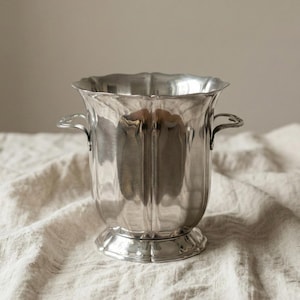 Könnte beinhalten: Eine silberfarbene Metallvase mit geriffeltem Korpus und gewelltem Rand. Die Vase hat zwei kleine Griffe und steht auf einem runden Sockel. Sie wird auf einem strukturierten, cremefarbenen Stoff präsentiert. Höhe ca. 20 cm.