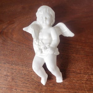 Könnte beinhalten: Eine weiße Porzellan-Cherub-Figur mit ausgebreiteten Flügeln und einem drapierten Tuch. Der Cherub hält eine Traube. Die Figur steht auf einer Holzoberfläche.