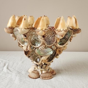 Puede incluir: Un cuenco decorativo de conchas marinas con base de pedestal. El cuenco está adornado con varias conchas marinas, incluyendo conchas de abulón, y presenta un color beige claro. El cuenco se utiliza probablemente con fines decorativos.