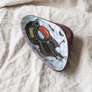 May include: A small, triangular ceramic dish with a dark maroon exterior and a white interior. The centre features a black graphic design with a red oval, a light blue oval, and a yellow rectangle, surrounded by white painted cloud-like shapes.