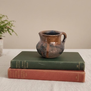 Puede incluir: Una pequeña jarra de cerámica marrón y azul, de estilo rústico, con asa, se encuentra sobre dos libros antiguos. La jarra tiene un cuerpo redondeado y un interior azul oscuro. Una planta verde en maceta está en el fondo.