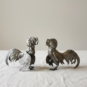 Könnte beinhalten: Zwei silberfarbene Hahnfiguren stehen sich gegenüber. Die Hähne haben detaillierte Federn, markante Kämme und gebogene Schwänze. Sie stehen auf einer strukturierten, hellen Oberfläche vor einem neutralen Hintergrund. Die Figuren sind dekorativ.
