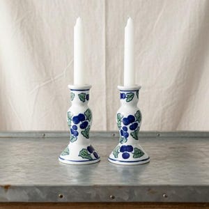 Può includere: Due candelabri in ceramica bianca con disegni di uva blu e foglie verdi. Ogni portacandela ha una candela bianca. I portacandele sono su una superficie metallica con una base in legno. Lo sfondo è un tessuto bianco.