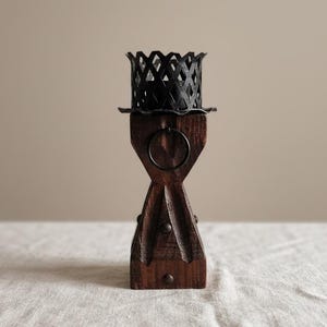 May include: A dark brown wooden candle holder featuring a tapered, hourglass-shaped base with vertical grooves and a small metal ring detail. The top is crowned with a black metal lattice cup designed to hold a candle securely in place.