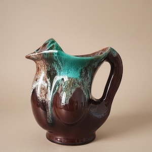 May include: A ceramic pitcher featuring a glossy dark brown base with a decorative drip glaze effect in vibrant turquoise and white cascading from the rim. The vessel has a rounded body, a wide pouring spout, and a curved handle on the side.