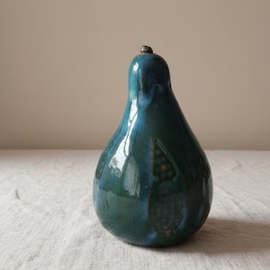 Puede incluir: Un objeto decorativo de cerámica con forma de pera, con un esmalte brillante de color verde-azulado. La pera tiene un pequeño tallo marrón oscuro y una forma lisa y redondeada. La superficie refleja la luz, resaltando las variaciones de color. El objeto está sobre una superficie neutra.