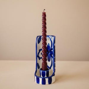Op de afbeelding: Een keramische kandelaar met een gebogen achterpaneel voorzien van blauwe handgeschilderde bloemmotieven en verticale blauwe strepen op de voet. Een enkele donkerbordeauxrode gedraaide taps toelopende kaars staat in de houder.