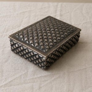 Pode incluir: Uma caixa retangular de metal com um acabamento escuro e oxidado. A tampa apresenta um padrão de grelha em relevo com formas de diamante, enquanto os lados da base são decorados com um design texturizado de cristas diagonais.