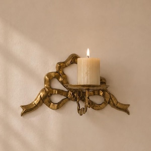 May include: A gold-toned wall-mounted candle holder shaped like a decorative ribbon bow. The metal fixture features a circular platform supporting a single, thick, cream-colored pillar candle with a lit flame, creating a warm, focused glow.