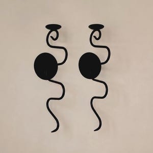 Puede incluir: Un par de candelabros de pared de metal negro con un diseño vertical, ondulado y serpenteante. Cada candelabro incluye una placa trasera circular para el montaje y una pequeña plataforma circular plana en la parte superior para una vela.