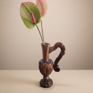 May include: A dark brown, carved wooden vase with a curved handle displays two anthurium flowers. One flower is green with pink veins, the other is pink. The vase has detailed carvings on the base and body.
