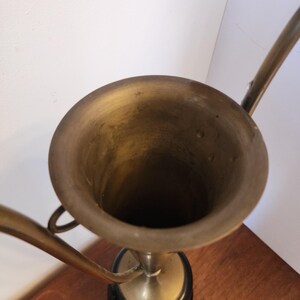 Pode incluir: Uma ta&ccedil;a de trof&eacute;u em lat&atilde;o com uma abertura circular larga e duas al&ccedil;as curvas que se estendem pelos lados. A ta&ccedil;a est&aacute; montada sobre uma base circular preta s&oacute;lida, exibindo um acabamento met&aacute;lico com sinais vis&iacute;veis de idade e p&aacute;tina.