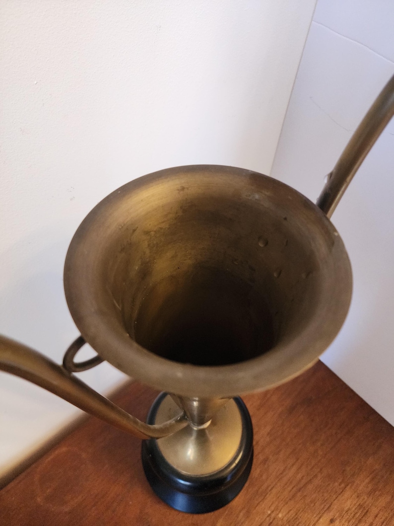Pode incluir: Uma ta&ccedil;a de trof&eacute;u em lat&atilde;o com uma abertura circular larga e duas al&ccedil;as curvas que se estendem pelos lados. A ta&ccedil;a est&aacute; montada sobre uma base circular preta s&oacute;lida, exibindo um acabamento met&aacute;lico com sinais vis&iacute;veis de idade e p&aacute;tina.