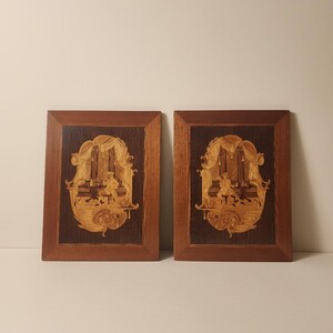 Op de afbeelding: Een paar rechthoekige houten wandborden met ingewikkeld inlegwerk. Elk bord toont een tafereel van lichtgekleurd hout met een persoon zittend aan een tafel in een gedrapeerd interieur, tegen een achtergrond van donkerbruin hout.