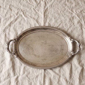 Op de afbeelding: Een ovaal zilverkleurig dienblad met een glad, reflecterend oppervlak en een decoratieve kralenrand. Het dienblad is voorzien van twee sierlijke, gebogen handgrepen aan weerszijden, elk gedetailleerd met ingewikkeld krulwerk en reliëfpatronen.