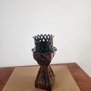 May include: A candle holder featuring a dark brown, four-sided wooden base with metal studs and a black metal top with a decorative lattice pattern. A small black metal ring is attached to the upper section of the wooden pillar structure.