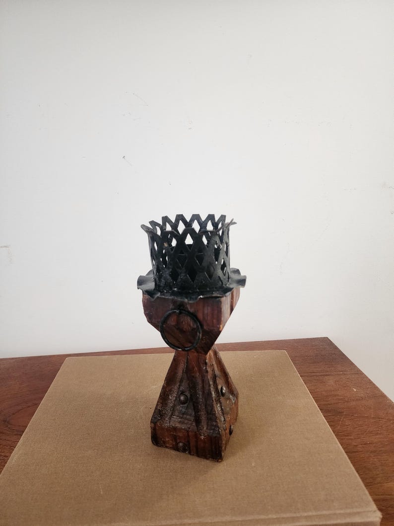 May include: A candle holder featuring a dark brown, four-sided wooden base with metal studs and a black metal top with a decorative lattice pattern. A small black metal ring is attached to the upper section of the wooden pillar structure.