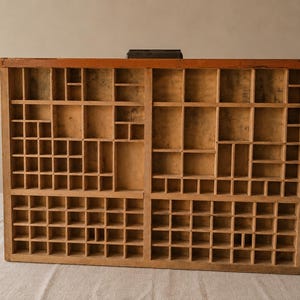 May include: A rectangular wooden printer's tray featuring a grid of numerous small, open compartments of varying sizes. The natural light-colored wood shows visible grain and texture, with a dark handle attached to the top center edge.